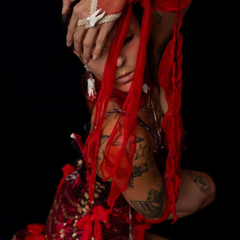 This image depicts the model LU crouched on the floor, body facing to the right with their head turned to the camera. Their hand is resting on their head while tassels from their glove trail over their face. 