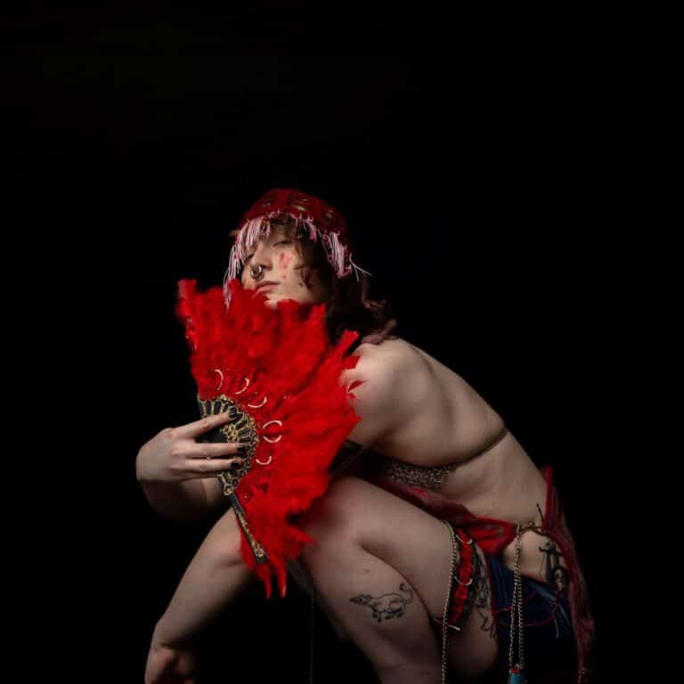 Cameron (model) squats on the floor facing the left with their head turned to camera, peering over a feather fan. 