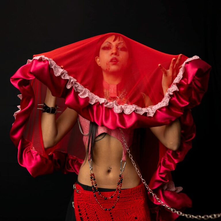 Vellatrix stands facing the camera, veil draped over their head. 