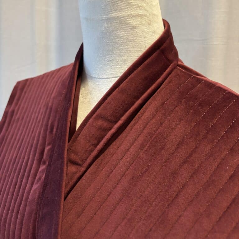 Close up of front of quilted fabric wearable device with large rectangular panels overlapping at the front. The fabric is soft plum coloured velvet. The garment is positioned on a mannequin in front of a curtain.