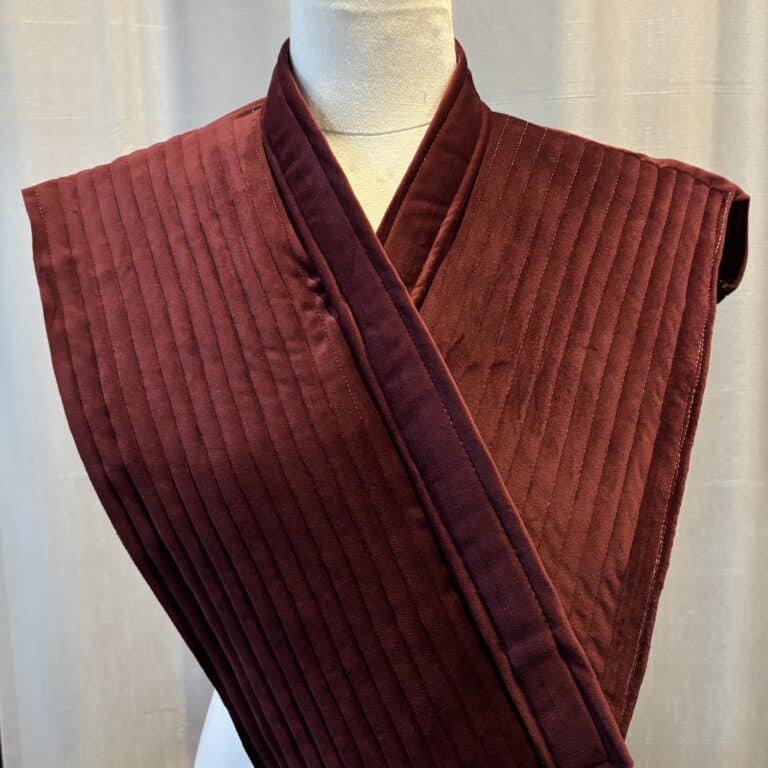 Quilted fabric wearable device with large rectangular panels overlapping at the front. The fabric is soft plum coloured velvet. The garment is positioned on a mannequin in front of a curtain.