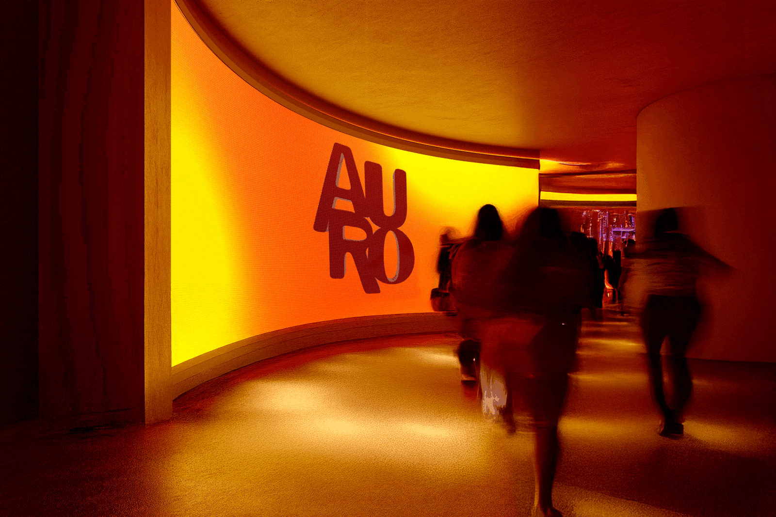 This image describes the ambience and nightclub of AURO. The Image captures the shutter speed of peoples shadows entering the warm moody entrance. the entrance has a large floor to wall LED screen featuring an illuminated gradient orange and sunset colour tones with the AURO logo rotating. 