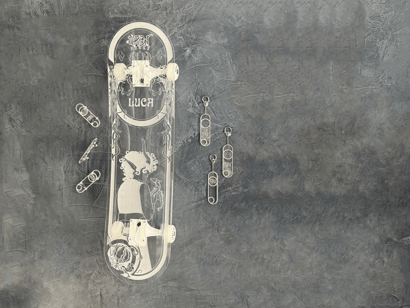 Clear acrylic skateboard with character art in art nouveau style along left-most rule of third line. To the left, three mini acrylic skateboards are scattered in a stack. On the right of the big skateboard are three keychains that are of mini skatedeck design (just with no wheels). Background is a grey painterly wall.