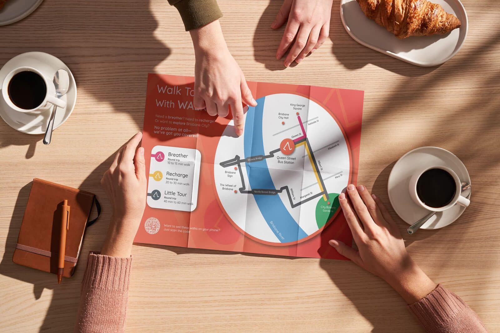 Two people exploring physical WALKKA maps placed on a café table, showing walking routes and community points of interest. The maps provide an accessible guide for users without smart devices, helping people discover local hidden gems and connect with their neighbourhood.